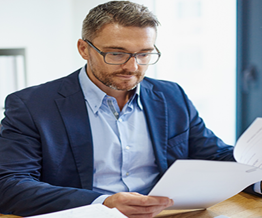 Image of a business man seated at a desk. He is wearing glasses and reviewing a lengthy document, which is secured by a paperclip