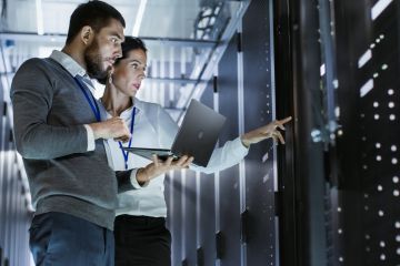 Two IT technicians standing in a server room in front of the racks reviewing and discussing something